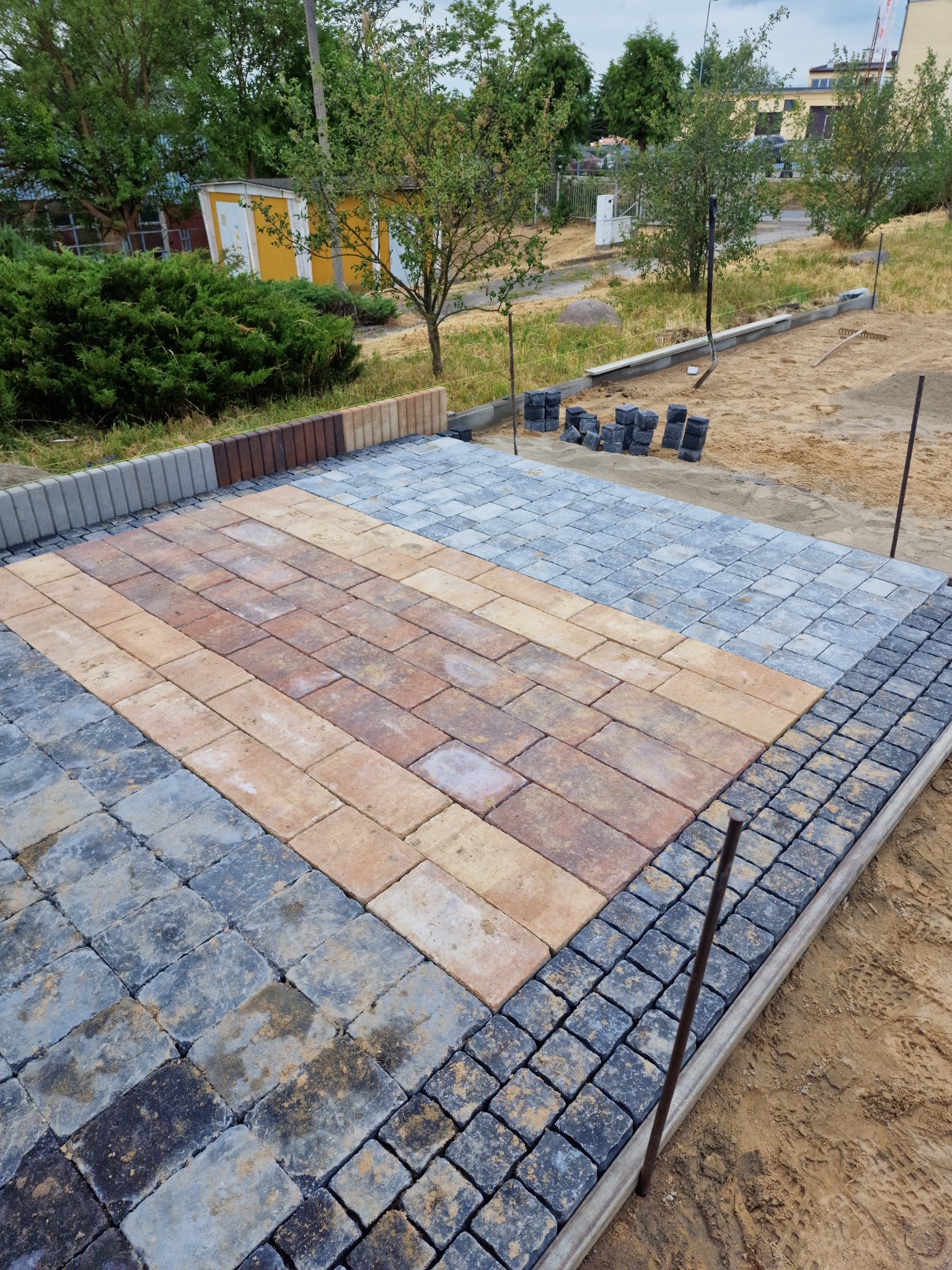 Fragment powierzchni z ułożoną kostką brukową w trzech różnych kolorach i wzorach, obrzeże z szarej kostki, widoczne narzędzia i materiały przygotowane do dalszych prac, w tle zieleń i budynek.