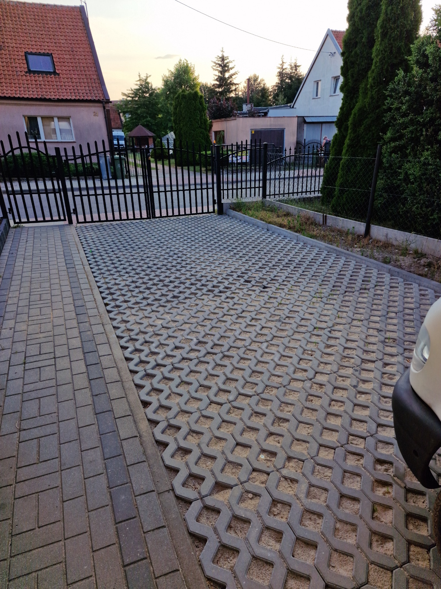 Szara ażurowa kostka brukowa na podjeździe, widok z góry na ułożoną nawierzchnię z widoczną bramą wjazdową i fragmentem samochodu.