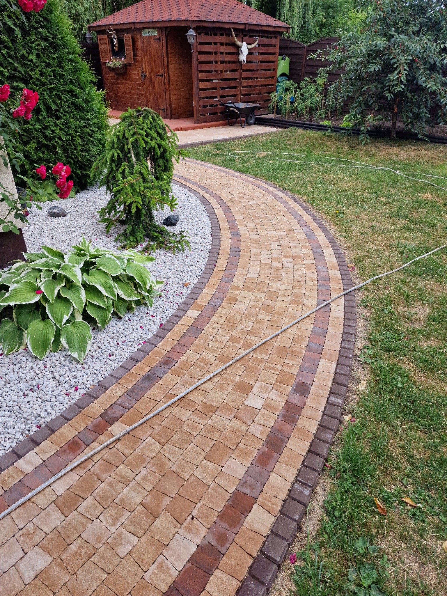 Zakrzywiony chodnik z kostki brukowej w odcieniach brązu i beżu, obrzeża z ciemniejszej kostki, obok rabata z białym kamieniem i roślinami, w tle altana z dekoracją czaszki zwierzęcej.