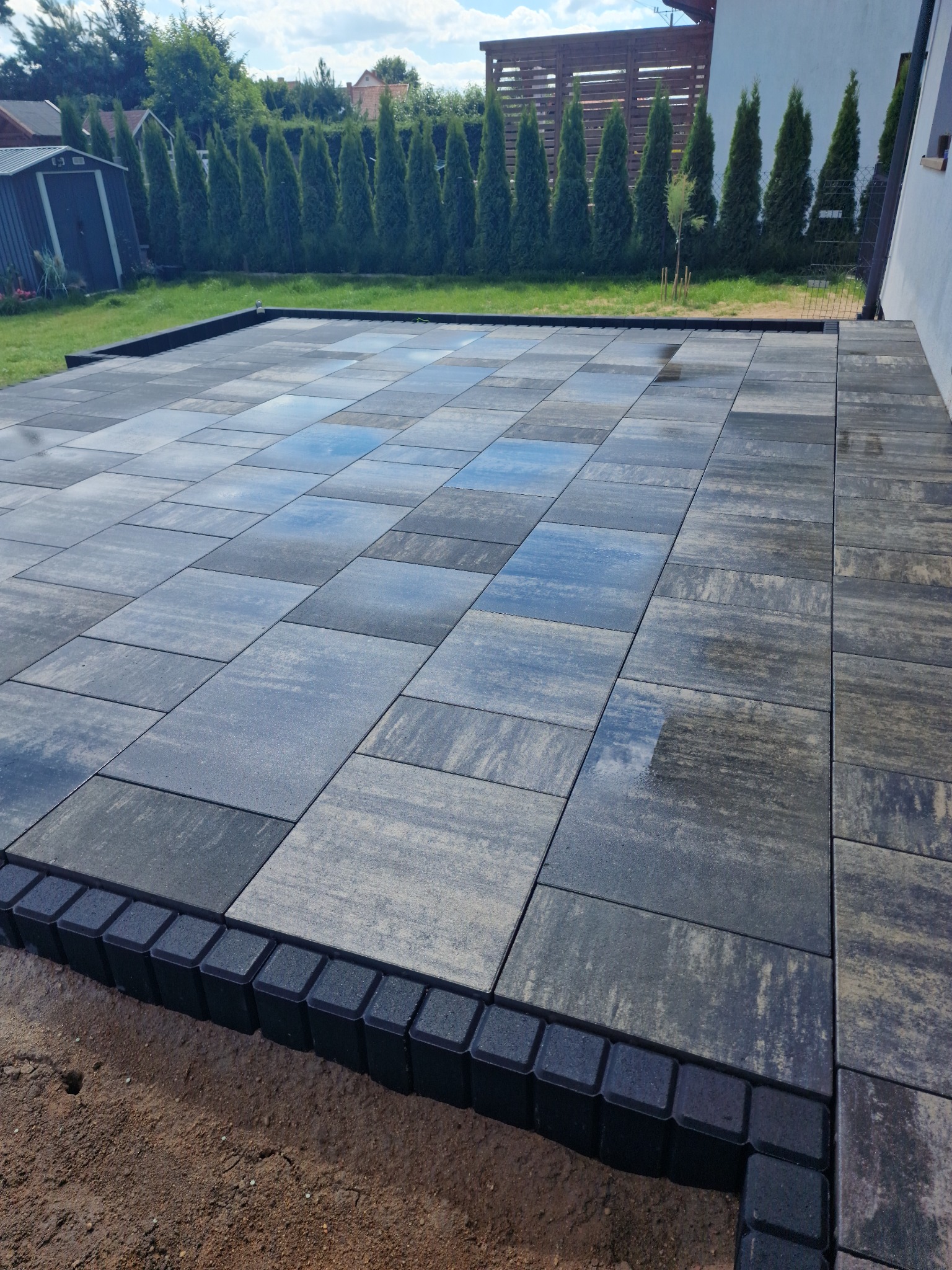 Ułożona kostka brukowa w odcieniach szarości i czerni, z widocznymi refleksami wody, zakończona czarnym obrzeżem, na tle trawnika i iglaków.