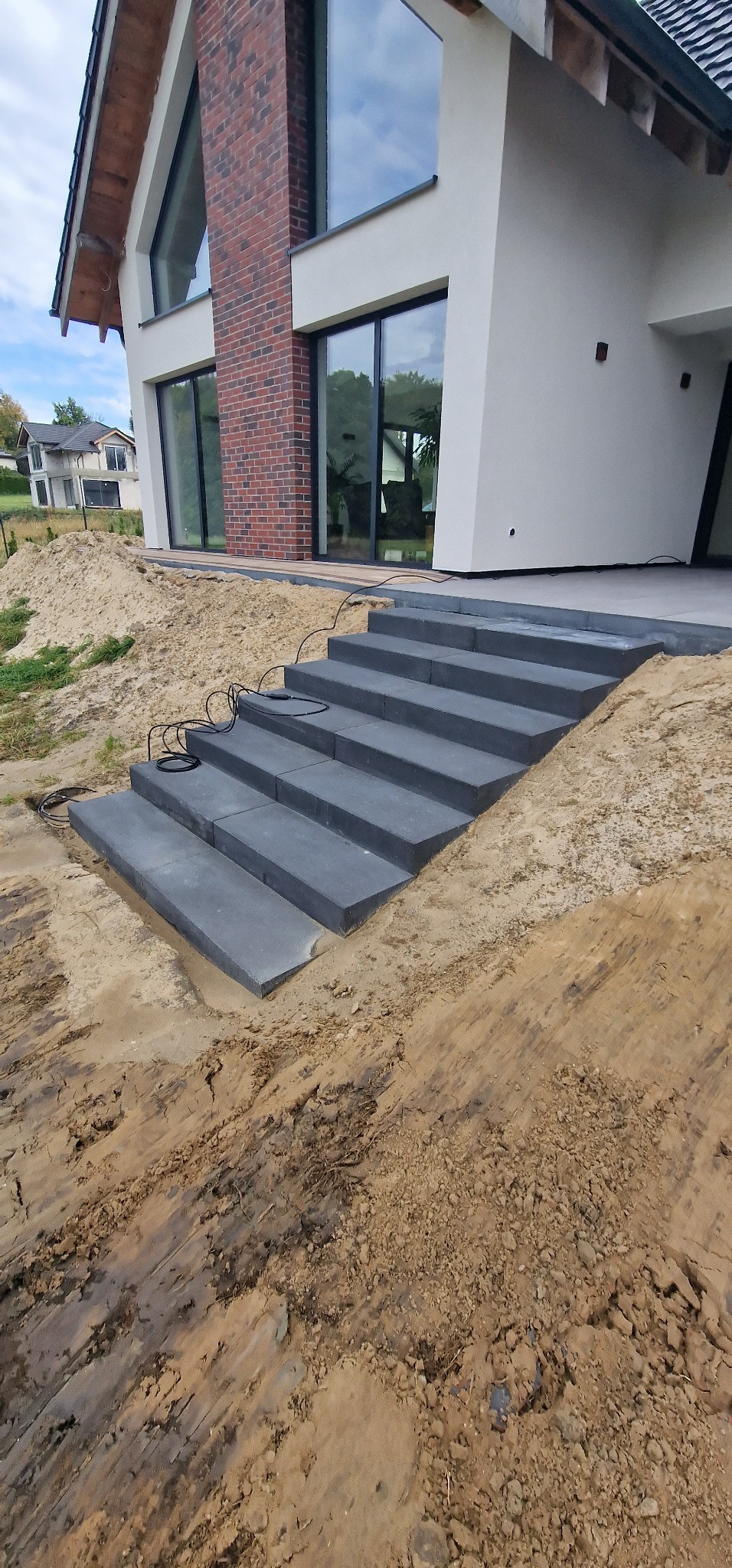 Schody z szarej kostki brukowej prowadzące do nowoczesnego domu z ceglanym kominem, widoczne ślady prac ziemnych w otoczeniu.