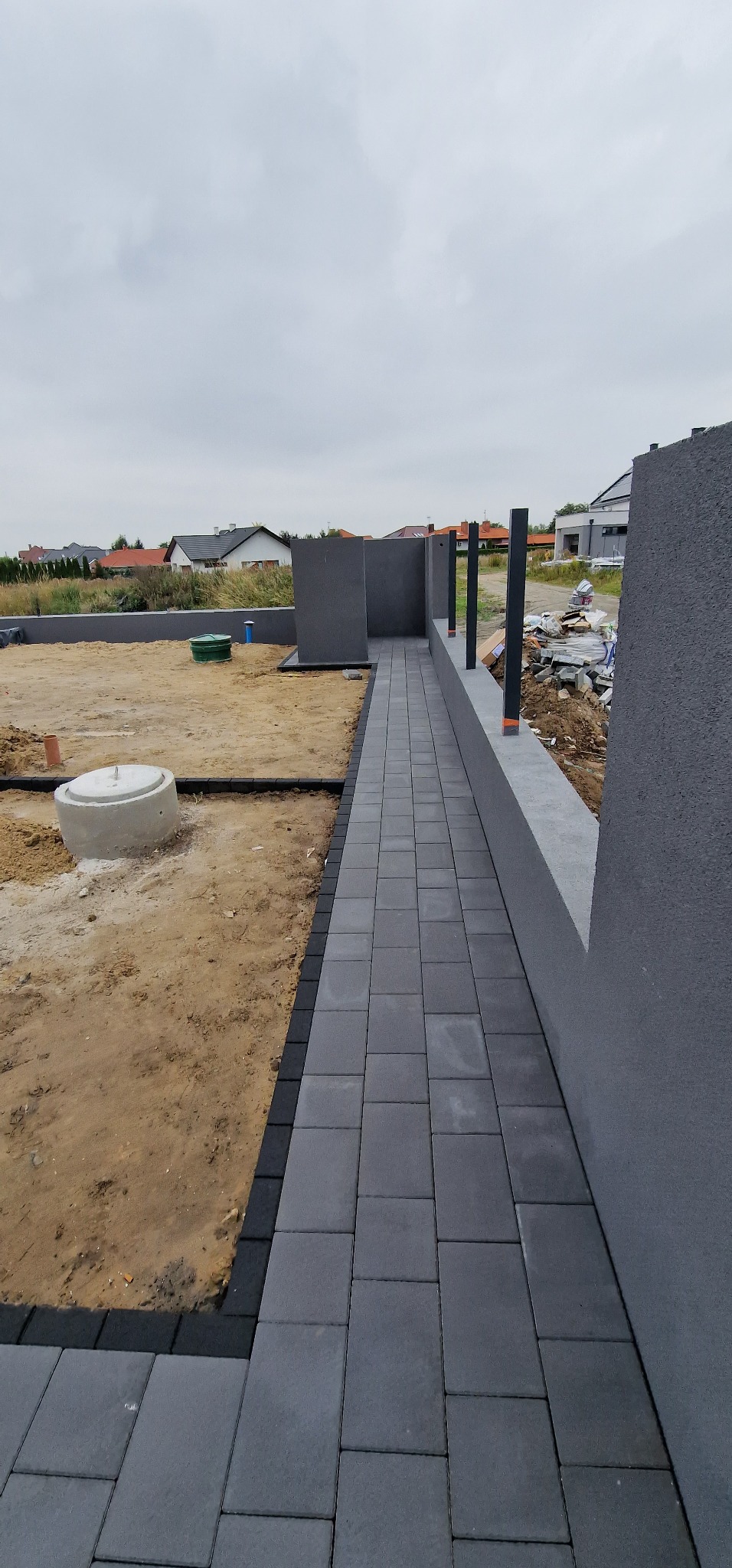 Ułożona ścieżka z szarych kostek brukowych prowadząca wzdłuż betonowego murku, z widocznymi słupkami ogrodzeniowymi w tle i niezabudowaną przestrzenią po lewej stronie z betonowym kręgiem.