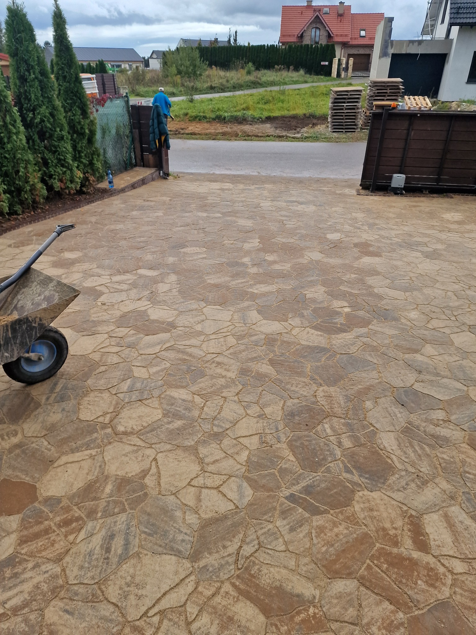 Wyłożony wzorzystą kostką brukową podjazd do domu z widoczną taczką z piaskiem, sugerującą trwające prace wykończeniowe.