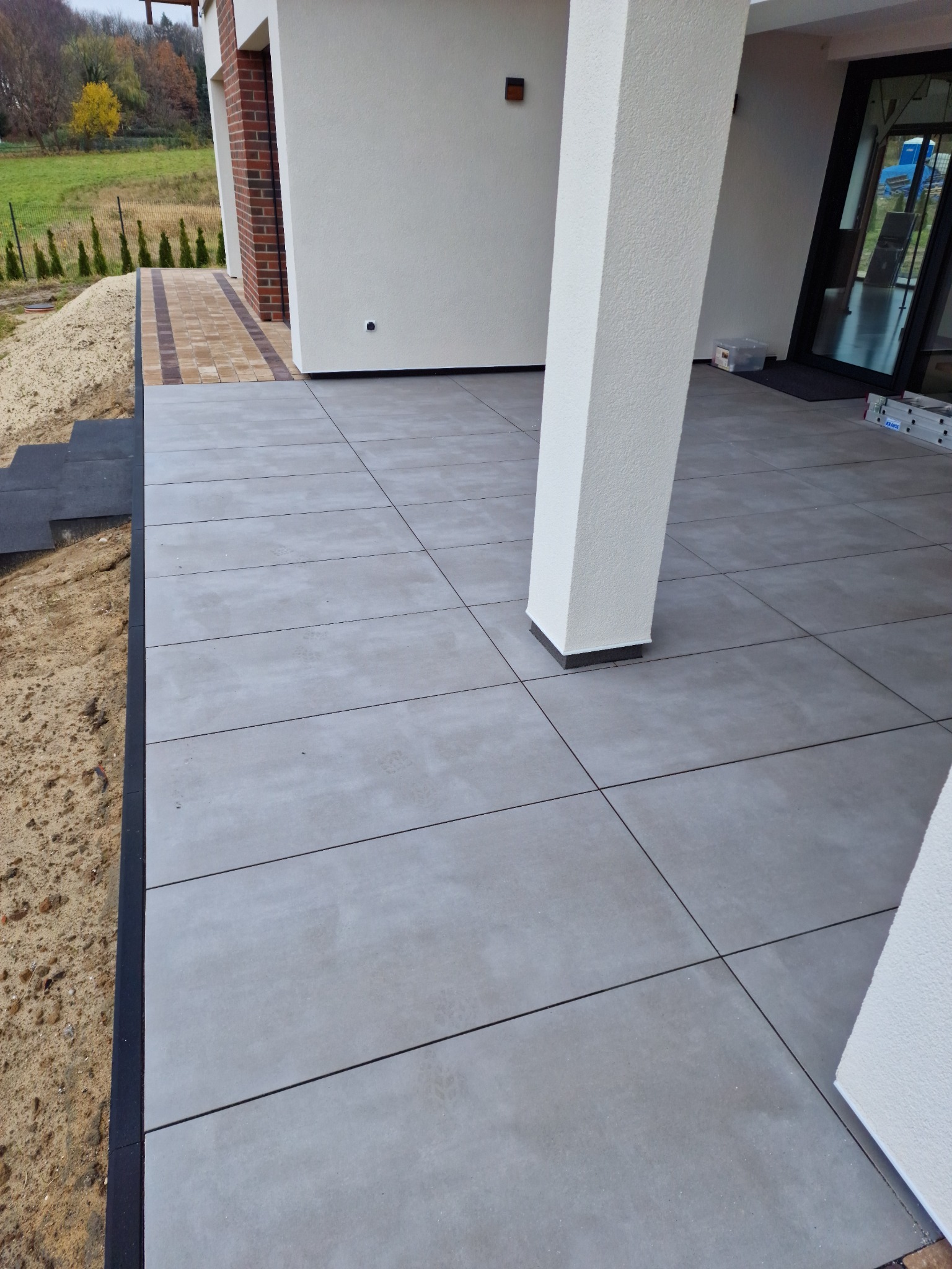 Szary taras wyłożony dużymi, prostokątnymi płytami, z widocznym słupem podtrzymującym zadaszenie i przygotowanym podłożem pod trawnik.