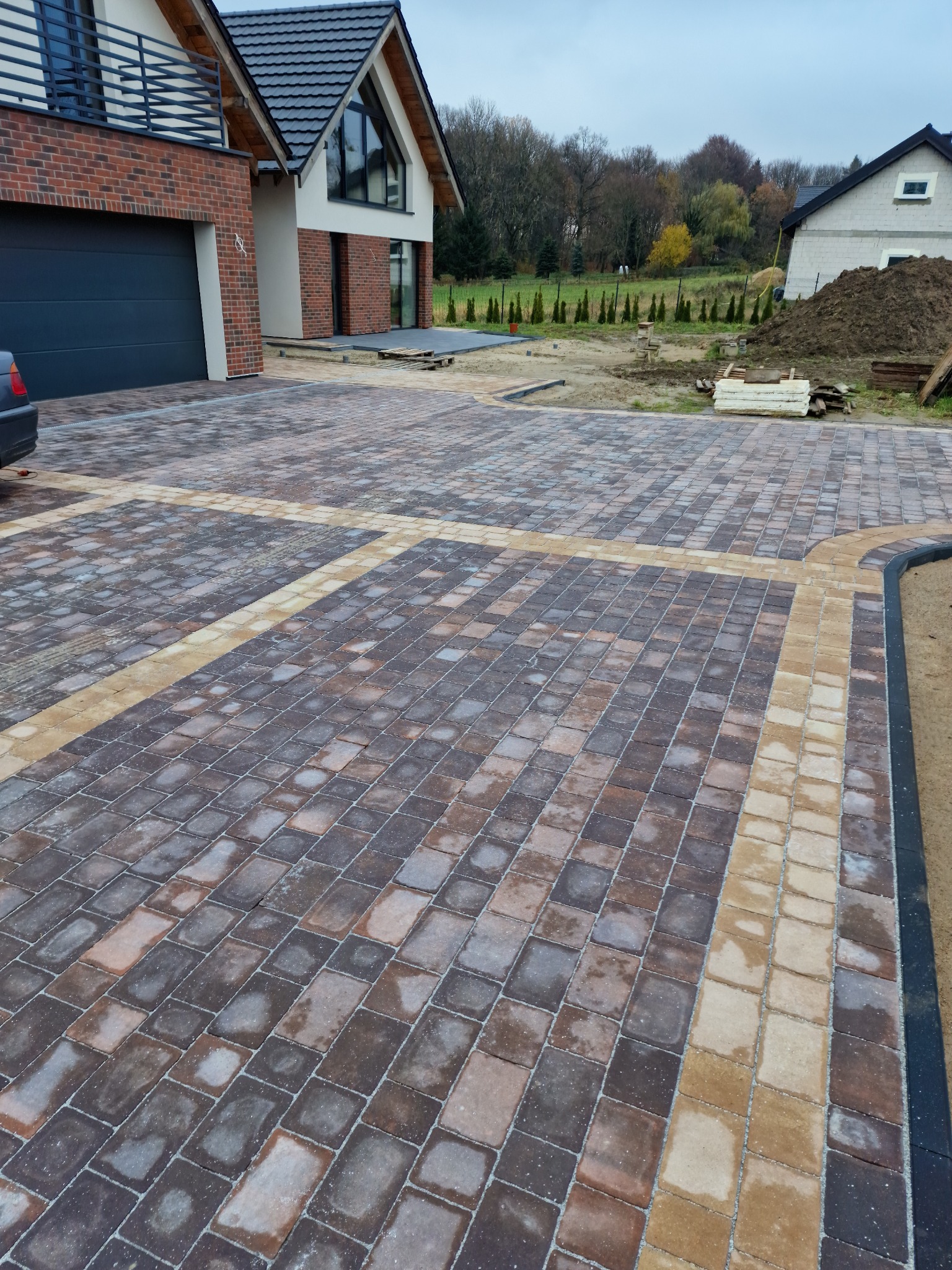 Ułożona kostka brukowa w dwóch odcieniach, tworząca wzór na podjeździe przed nowoczesnym domem z garażem i elementami ceglanymi.