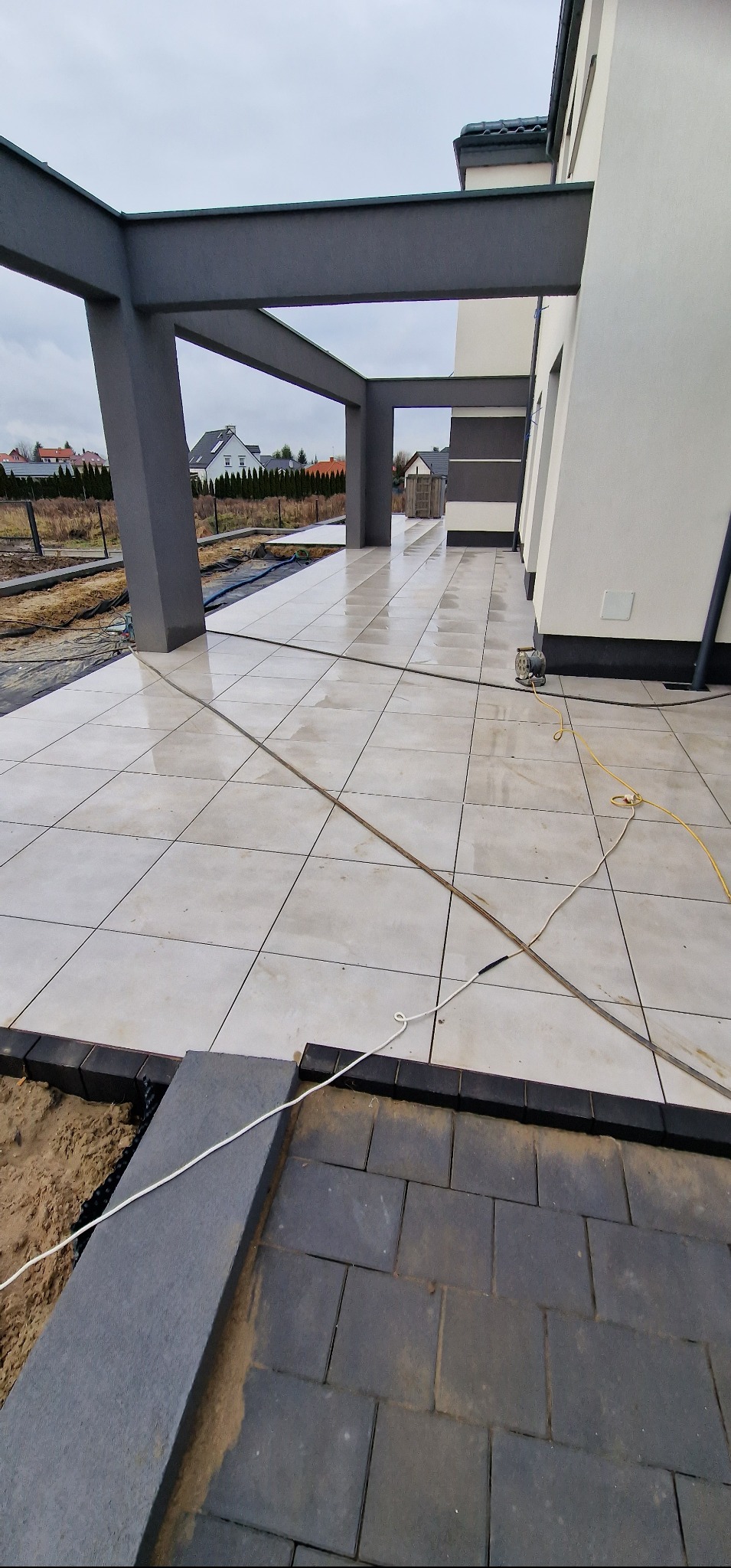 Wyłożony jasnymi płytami taras z szarą konstrukcją zadaszenia, obok trwają prace nad układaniem ciemnej kostki brukowej, widoczne narzędzia i piasek.