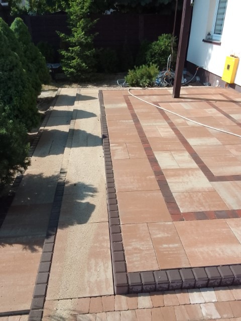 Nowo ułożona nawierzchnia z kostki brukowej w odcieniach beżu i brązu, z dekoracyjnym obramowaniem, prowadząca do tarasu przy domu z elementami drewnianej konstrukcji zadaszenia.