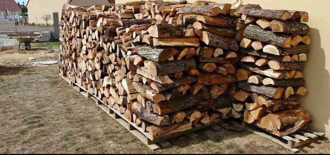 Rząd ułożonego drewna na palecie z widoczną korą, przygotowanego na opał, ustawiony na zewnątrz przy beżowej ścianie budynku.