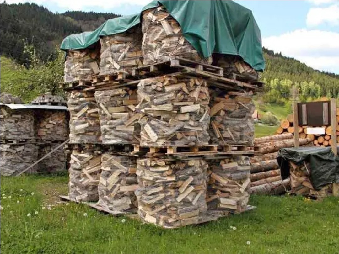 Stosy workowanego, pociętego drewna ułożone na paletach, zabezpieczone siatką i przykryte plandeką, z widocznym tłem górskim i składowanym drewnem w tle.