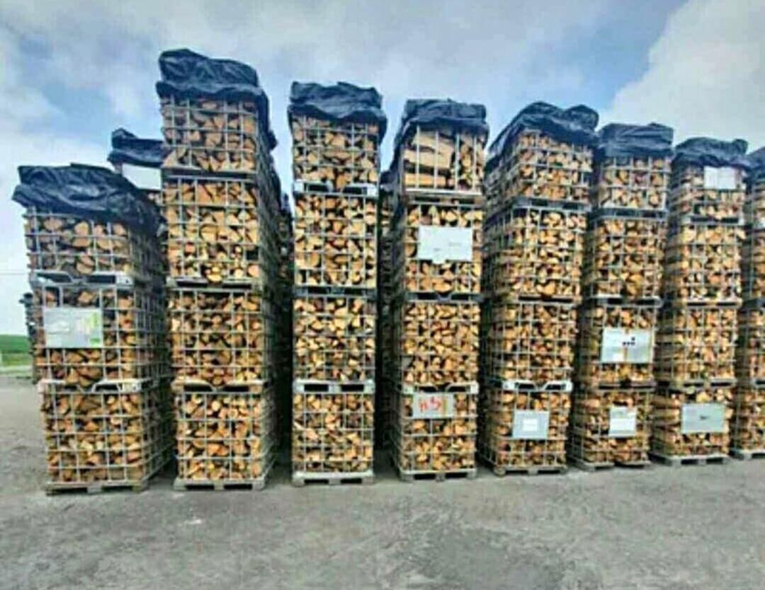 Rzędy metalowych koszy wypełnionych pociętym drewnem, częściowo przykrytych czarną folią, ustawione na betonowej powierzchni pod pochmurnym niebem.