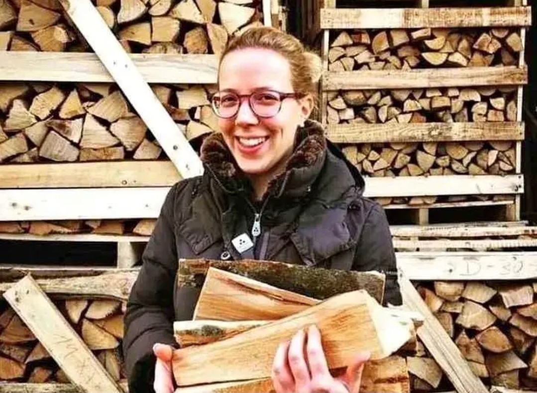 Uśmiechnięta kobieta w okularach i kurtce niesie naręcze drewna kominkowego na tle ułożonych stosów drewna w drewnianej konstrukcji.