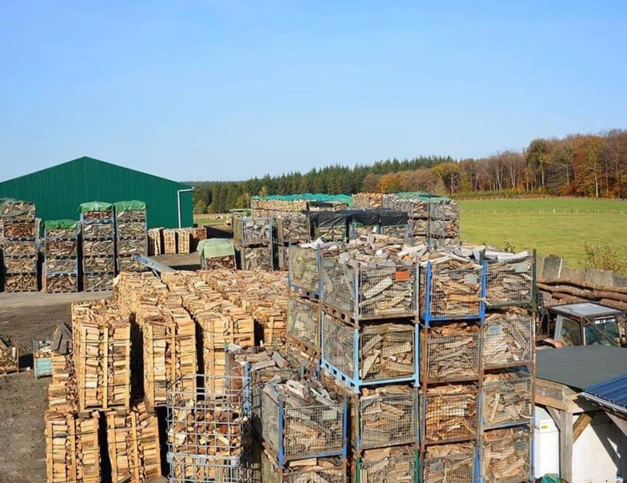 Stosy pociętego drewna ułożonego w metalowych koszach i na paletach, na placu składowym z zielonym budynkiem magazynowym w tle, widok z góry.