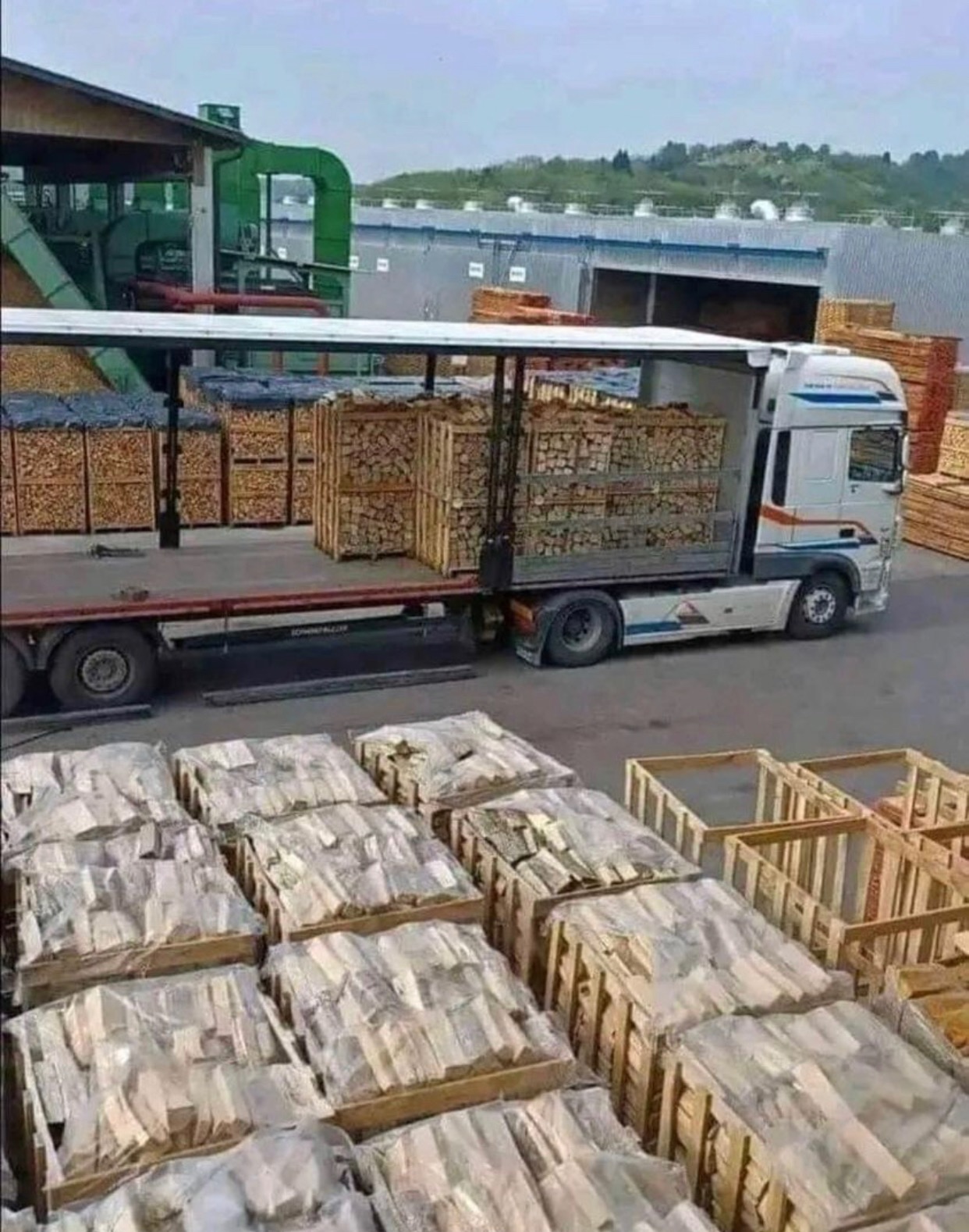 Ciężarówka z przyczepą załadowana drewnem pociętym na kawałki, ułożonym w drewnianych skrzyniach, obok stos skrzyń z drewnem w folii, na tle budynku przemysłowego i wzgórz.