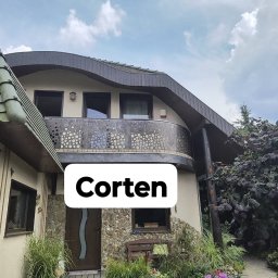 AT-TC - Dom z balkonem z balustradą ze stali corten o ażurowym wzorze, kamienne wykończenie elewacji, brązowe okna i drzwi, zielony dach. Widok z poziomu ogrodu.