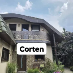 Dom z balkonem z balustradą ze stali corten o ażurowym wzorze, kamienne wykończenie elewacji, brązowe okna i drzwi, zielony dach. Widok z poziomu ogrodu.