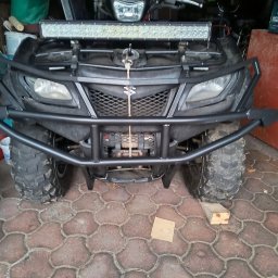 AT-TC - Czarny quad Suzuki z zamontowanym stalowym orurowaniem ochronnym i lampą LED. Widok z przodu, na pierwszym planie kostka brukowa.