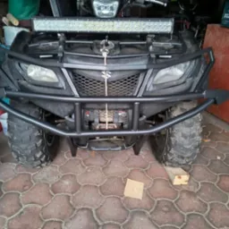 Czarny quad Suzuki z zamontowanym stalowym orurowaniem ochronnym i lampą LED. Widok z przodu, na pierwszym planie kostka brukowa.