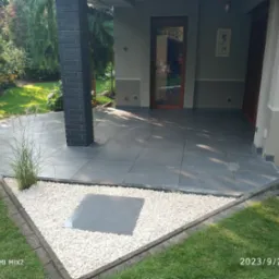 Nowoczesny taras z szarych płyt, obrzeża z białego kamienia i trawy. Ciemna kolumna z cegły, drzwi wejściowe w tle. Ujęcie z poziomu trawnika.