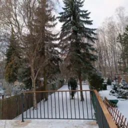 Zimowy krajobraz ogrodu widziany z tarasu z metalową balustradą o drewnianych poręczach, śnieg pokrywa drzewa i ziemię.