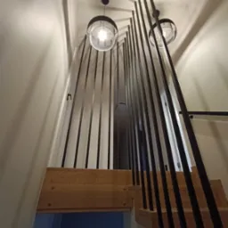 Nowoczesne schody drewniane z czarną balustradą z pionowych prętów, widok z dołu w kierunku sufitu z dwoma lampami.