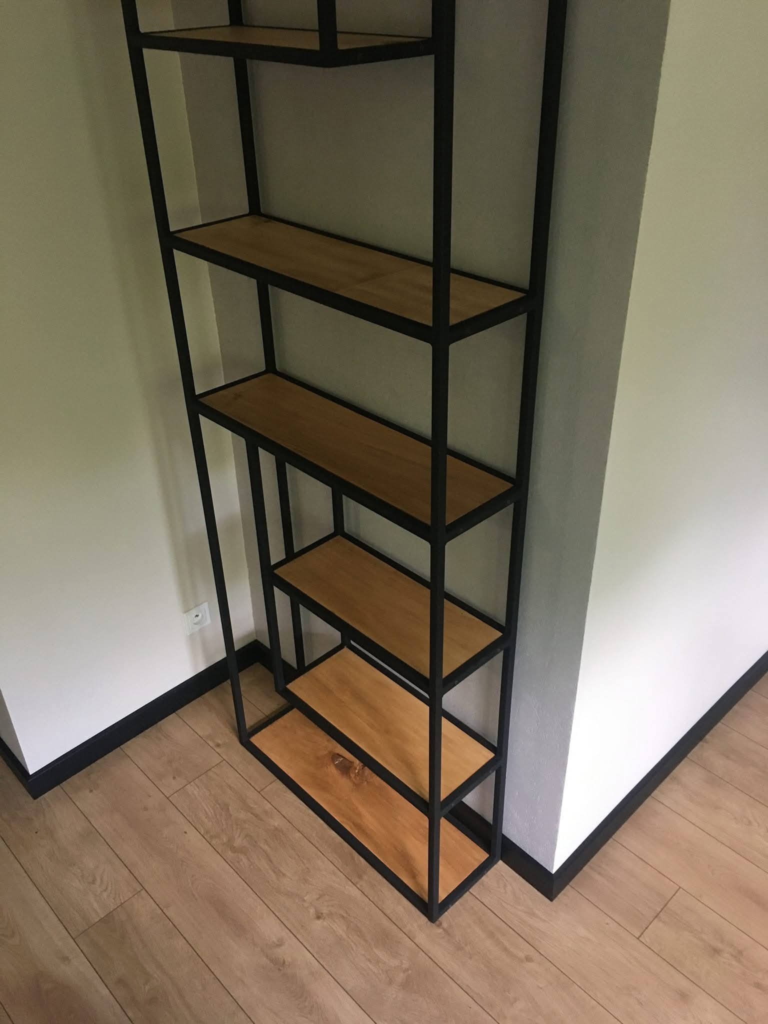 Regał loftowy z czarną metalową ramą i drewnianymi półkami, ustawiony w rogu pokoju z drewnianą podłogą i białymi ścianami, widoczny fragment gniazdka elektrycznego.