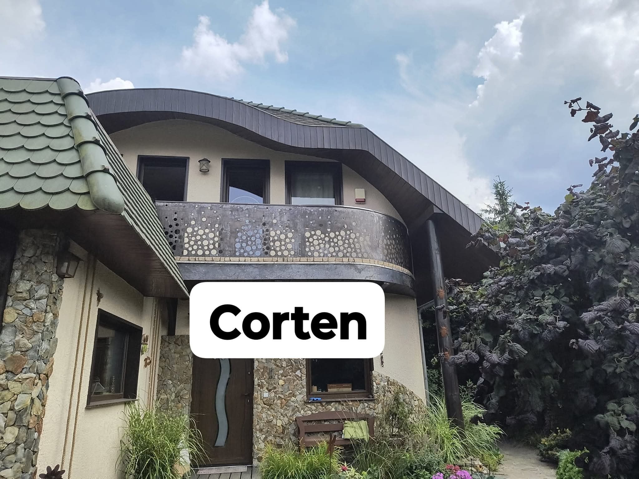 Dom z balkonem z balustradą ze stali corten o ażurowym wzorze, kamienne wykończenie elewacji, brązowe okna i drzwi, zielony dach. Widok z poziomu ogrodu.