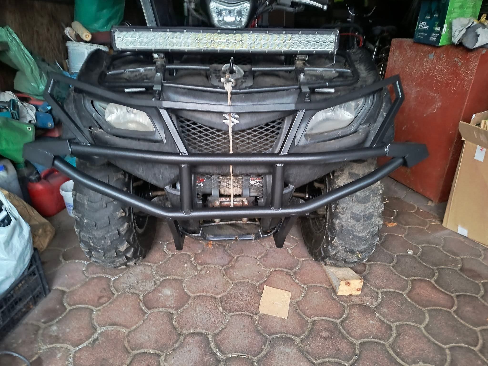 Czarny quad Suzuki z zamontowanym stalowym orurowaniem ochronnym i lampą LED. Widok z przodu, na pierwszym planie kostka brukowa.