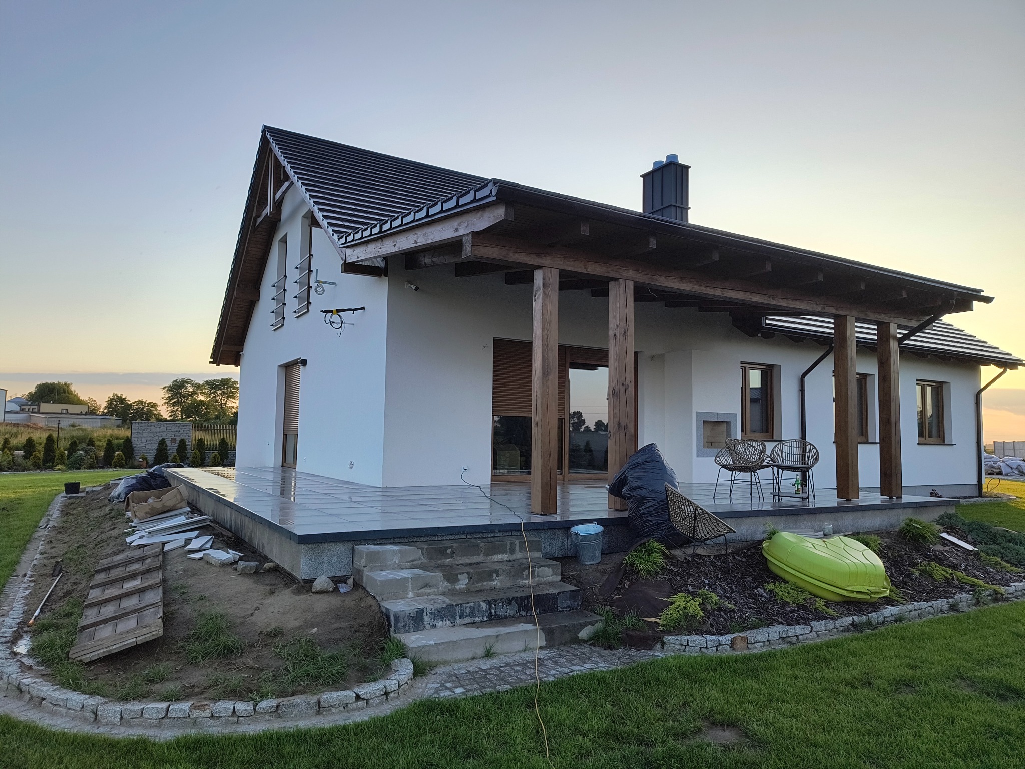 Nowoczesny dom z tarasem w trakcie wykańczania: szare płytki, drewniane kolumny podtrzymujące zadaszenie. Widoczne materiały budowlane i narzędzia.