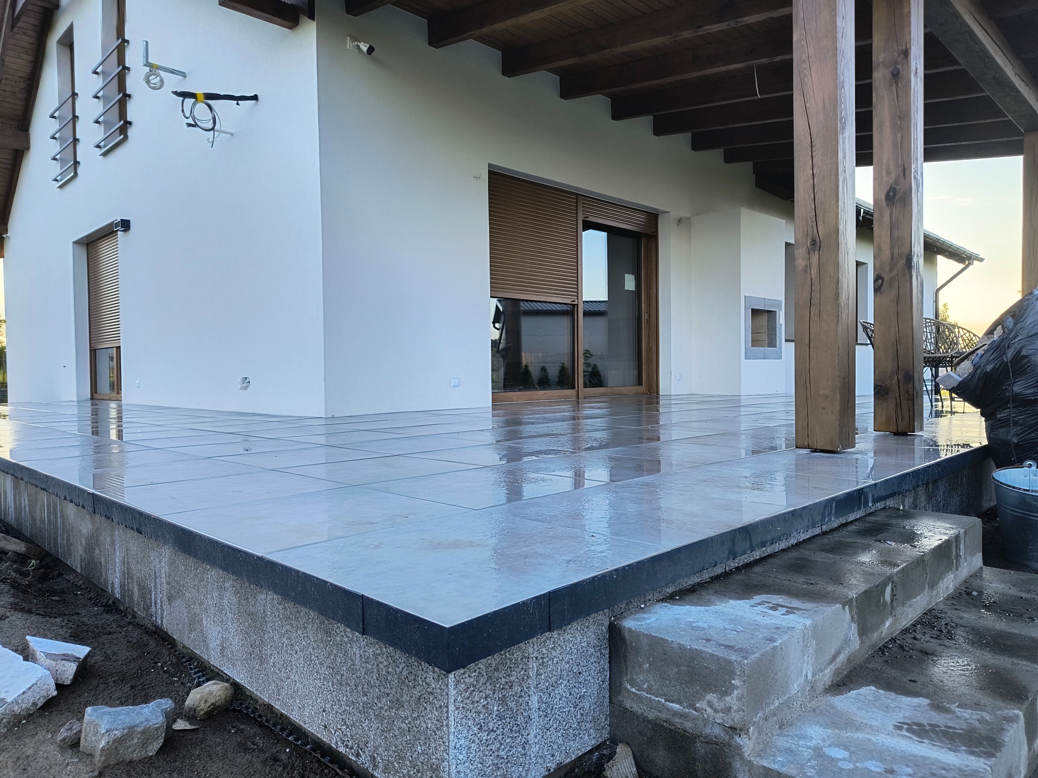 Nowoczesny taras betonowy z szarych płyt, wykończony czarną listwą. Widoczne drewniane słupy zadaszenia i fragment domu z roletami zewnętrznymi. Świeżo po deszczu.