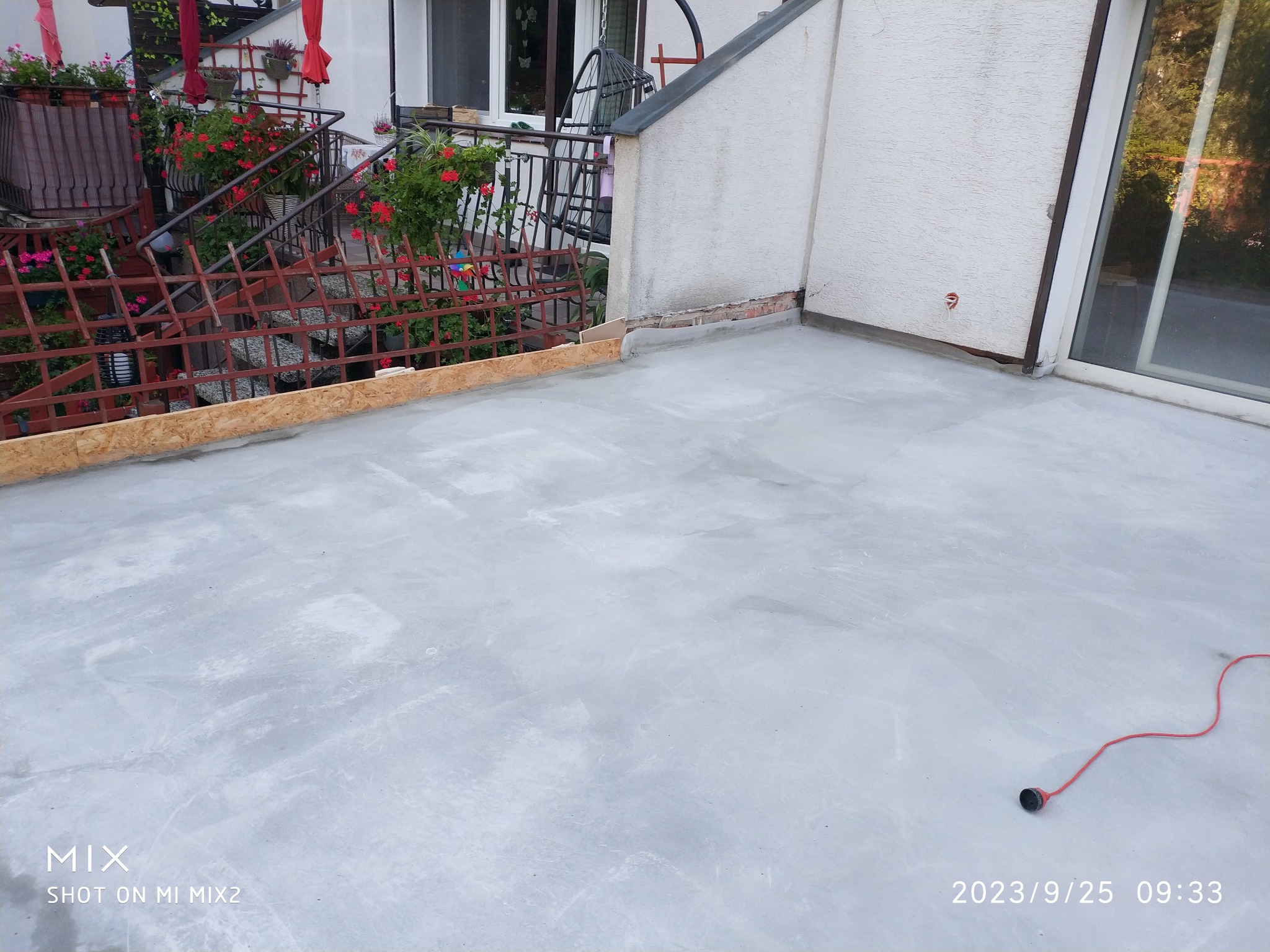 Świeżo wylany, szary taras betonowy z drewnianym obramowaniem i widoczną czerwoną wtyczką. W tle balustrady i kwiaty na balkonie.