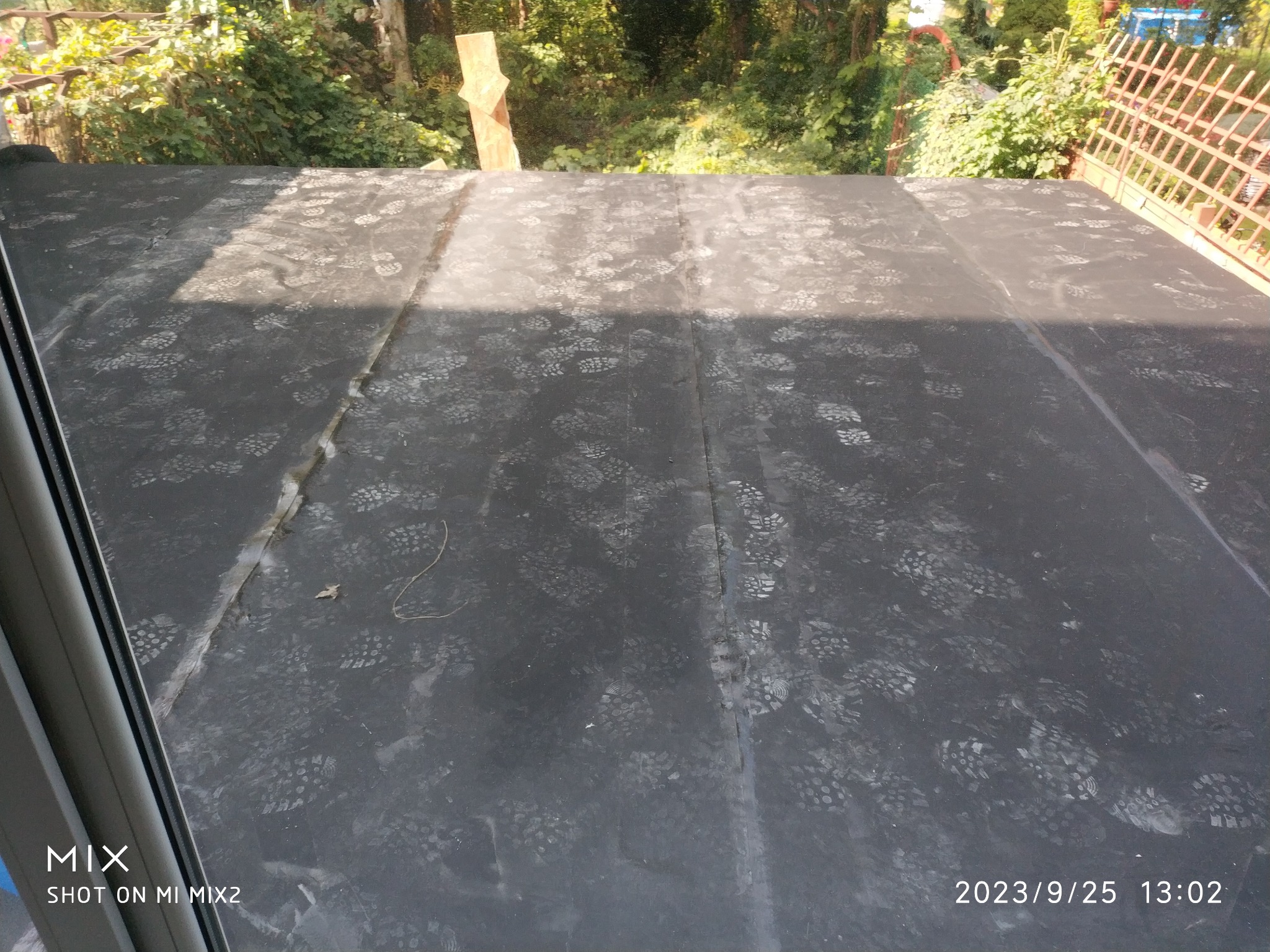 Czarna powierzchnia tarasu pokryta śladami stóp, widoczna zza okna, z zewnątrz zieleń drzew i fragment ażurowej konstrukcji.