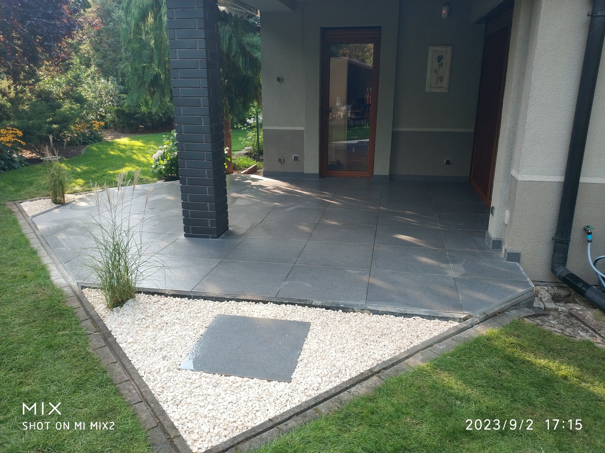 Nowoczesny taras z szarych płyt, obrzeża z białego kamienia i trawy. Ciemna kolumna z cegły, drzwi wejściowe w tle. Ujęcie z poziomu trawnika.