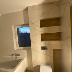 SkandyDam Damian Ziemba - Wykończona łazienka z wanną, toaletą i ścianą z marmurowym wzorem oraz wnękami. Widok na okno z zachodem słońca.