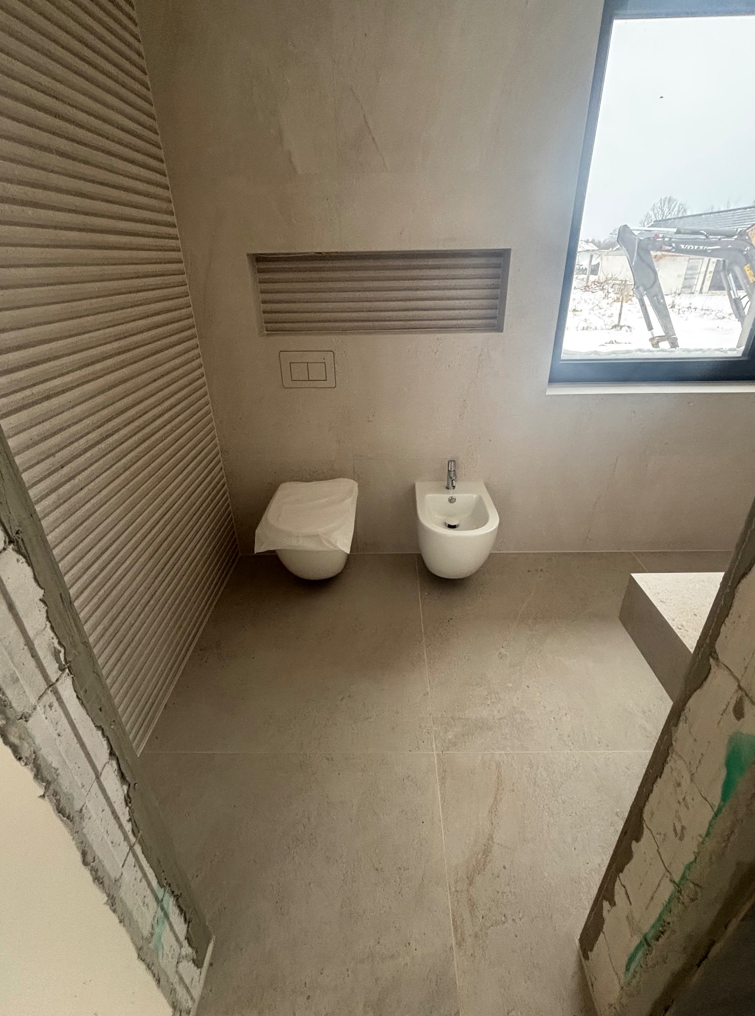 Jasna łazienka w trakcie wykańczania: bidet, toaleta z osłoną, okno z widokiem na zewnątrz, dekoracyjna ściana z poziomymi żłobieniami. Widoczne surowe wykończenia.