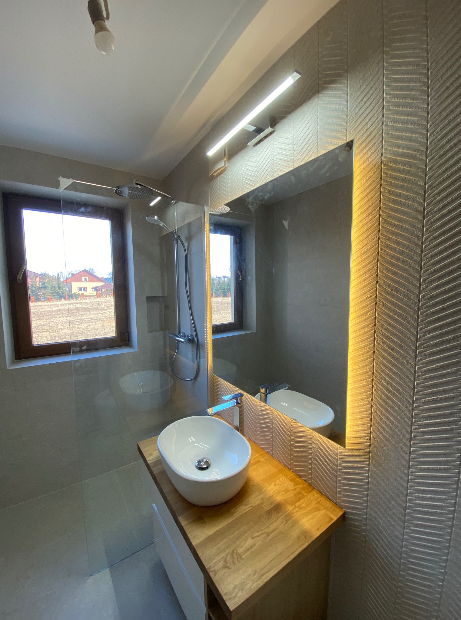 Nowoczesna łazienka z owalną umywalką na drewnianym blacie, podświetlanym lustrem i kabiną prysznicową. Na ścianie dekoracyjne płytki w jodełkę. Widok z okna na zewnątrz.