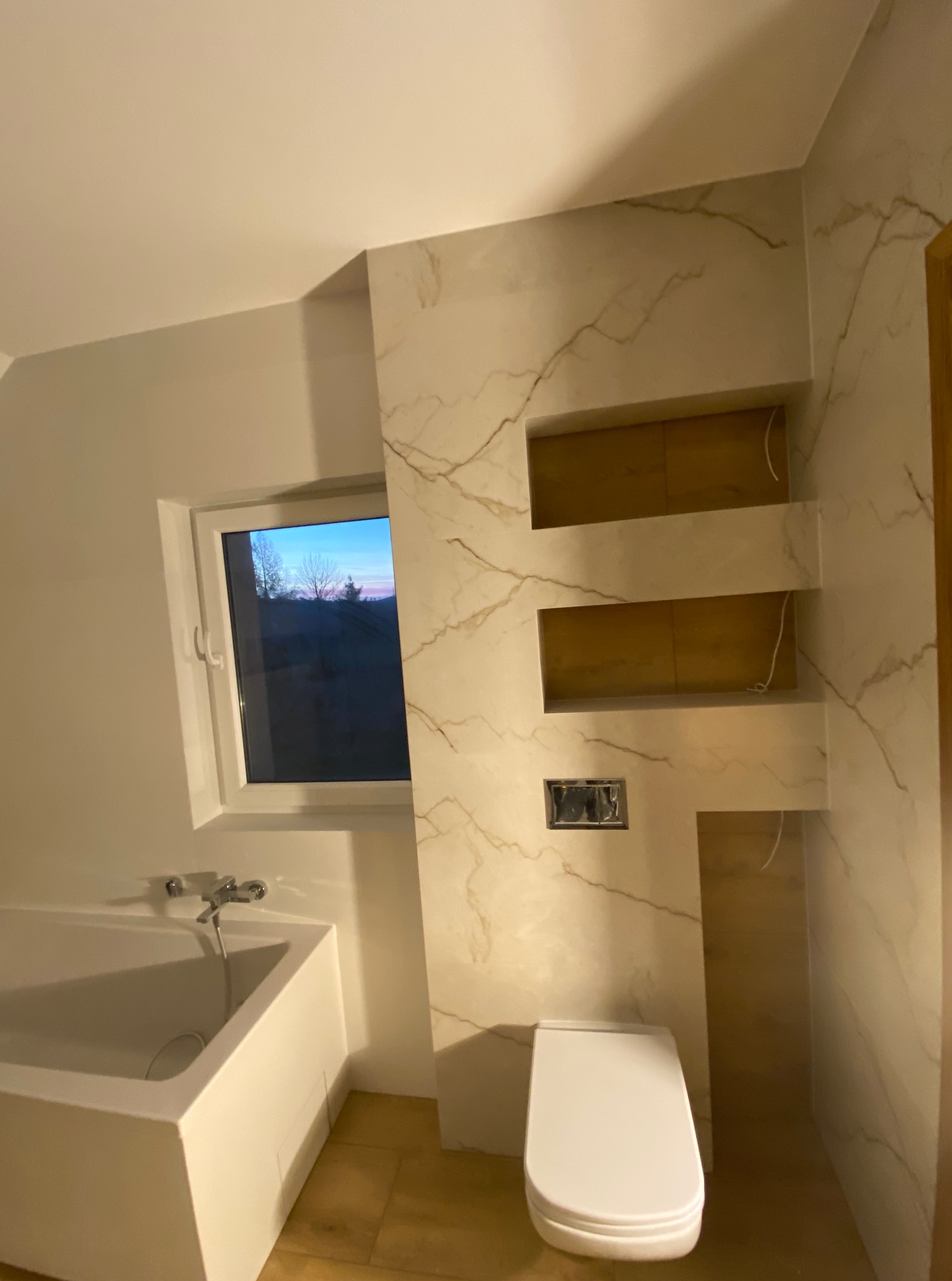 Wykończona łazienka z wanną, toaletą i ścianą z marmurowym wzorem oraz wnękami. Widok na okno z zachodem słońca.