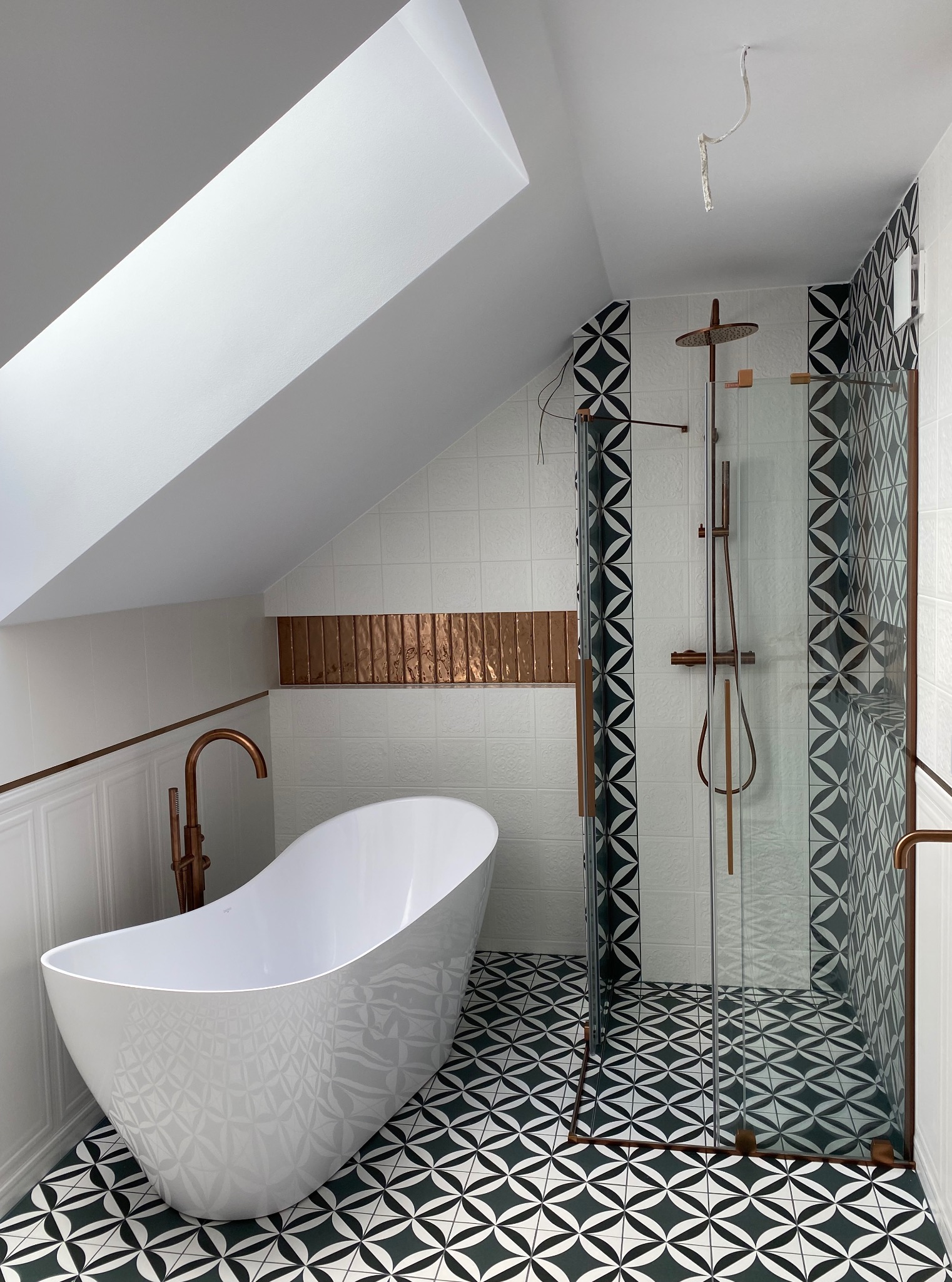 Nowoczesna łazienka na poddaszu z białą wanną wolnostojącą, kabiną prysznicową z miedzianymi elementami i wzorzystą podłogą w odcieniach bieli i zieleni.