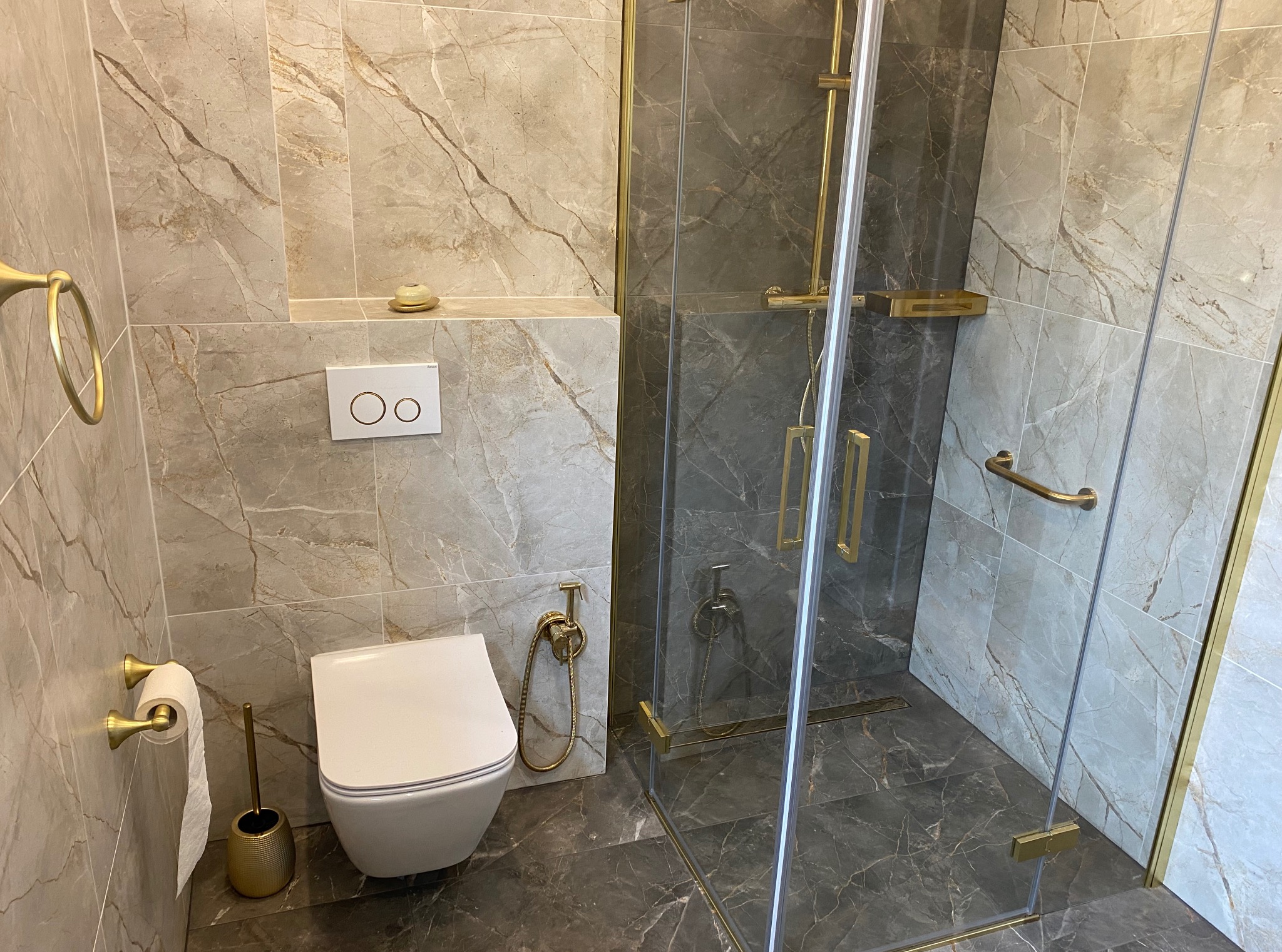 Wykończona łazienka z kabiną prysznicową, toaletą i złotymi akcentami. Płytki imitujące marmur na ścianach i podłodze. Nowoczesny design.