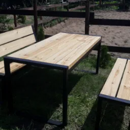 Zestaw mebli ogrodowych: stół i dwie ławki z jasnego drewna i czarnych metalowych profili, ustawiony na trawie, w tle ogrodzenie i piaskowe podłoże.