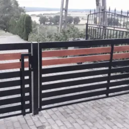 Nowoczesna brama wjazdowa z czarnych metalowych profili z poziomymi wstawkami z drewna, widoczna na tle kostki brukowej i zieleni ogrodu.