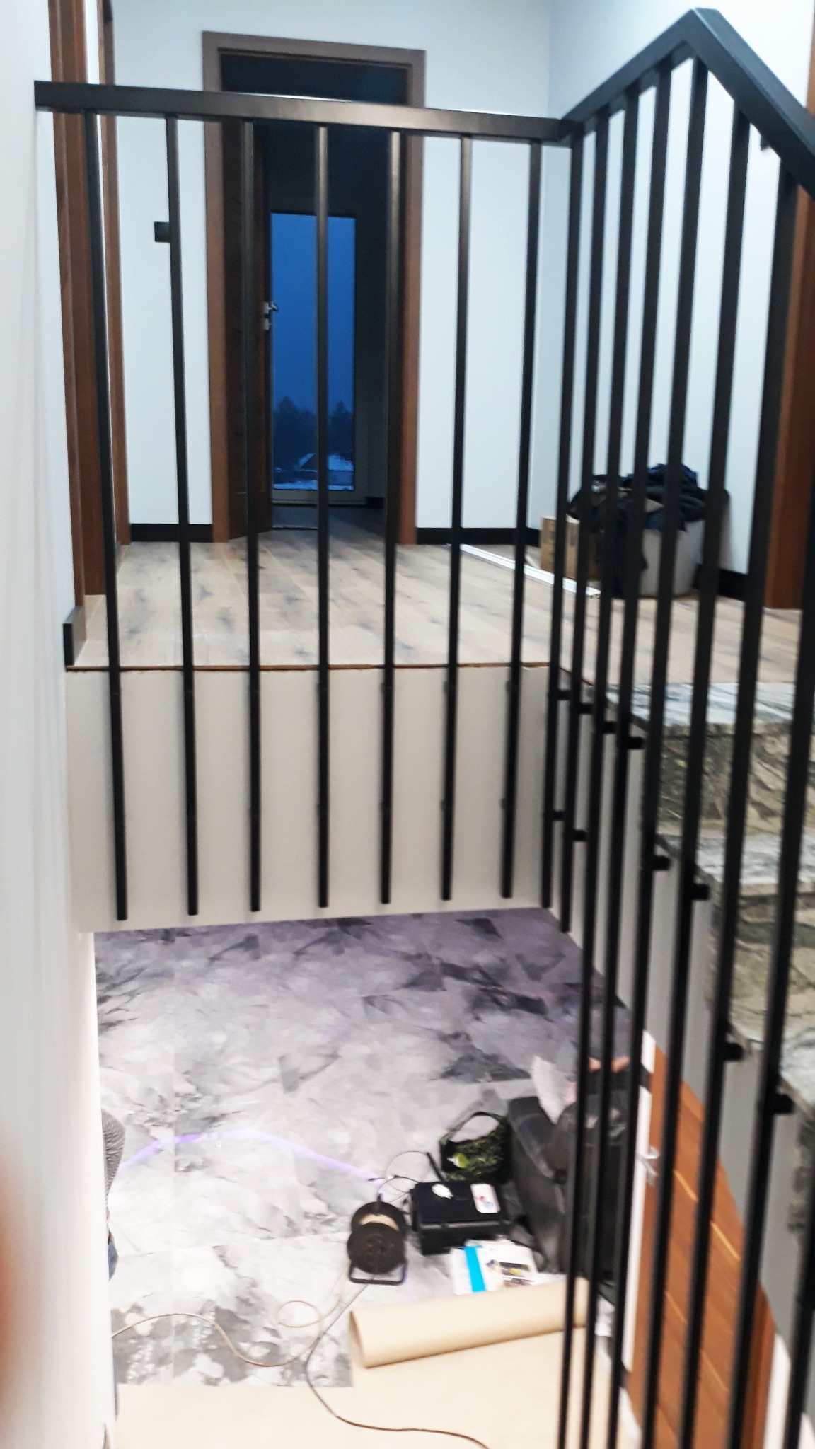 Częściowy widok na czarne, metalowe balustrady schodowe z pionowymi szczeblami, widoczne materiały budowlane i narzędzia na podłodze, w tle drzwi i okno z widokiem na zewnątrz.