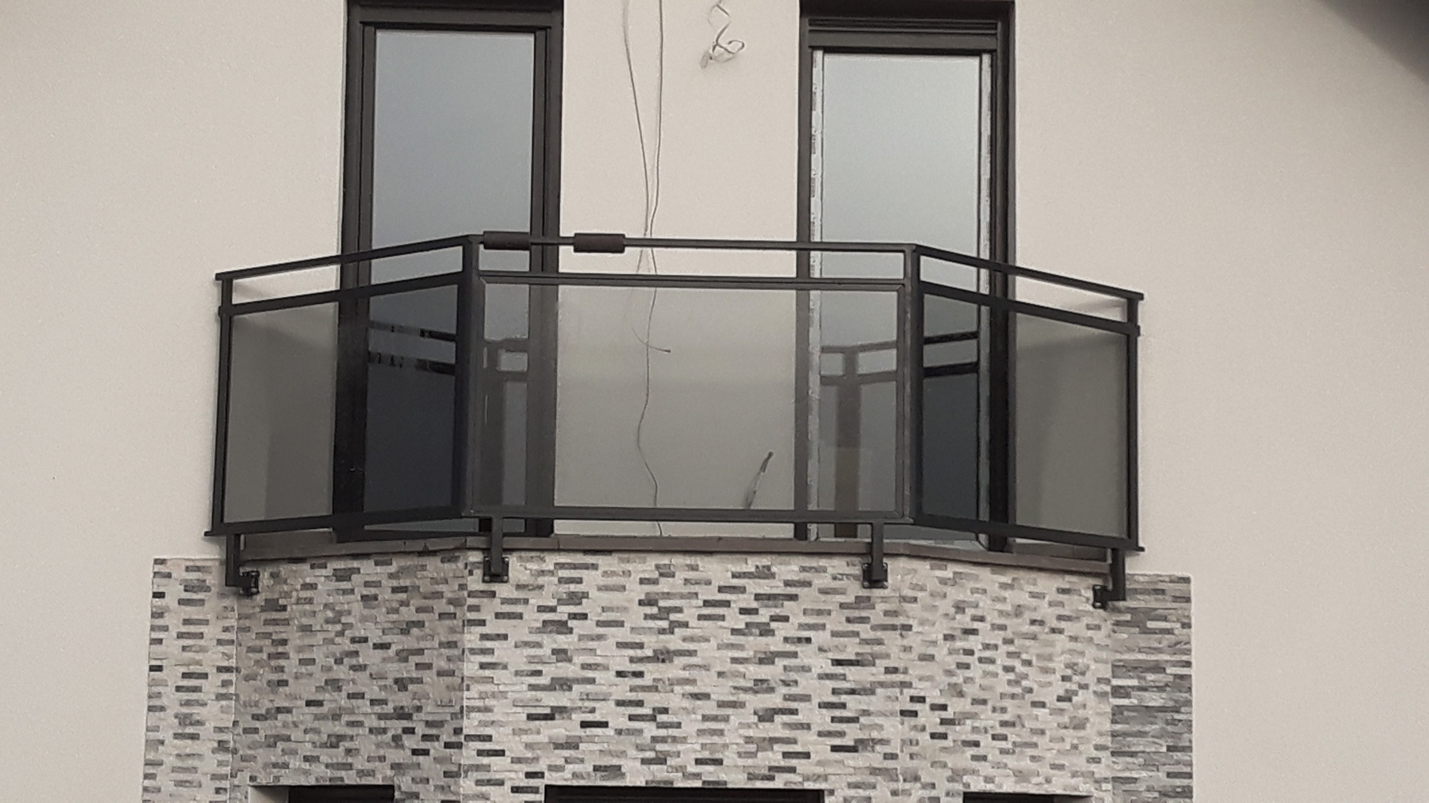 Nowoczesna balustrada balkonowa z czarną ramą i szarym szkłem, zamontowana na elewacji z dekoracyjnej cegły, widoczne przewody elektryczne sugerujące prace wykończeniowe.