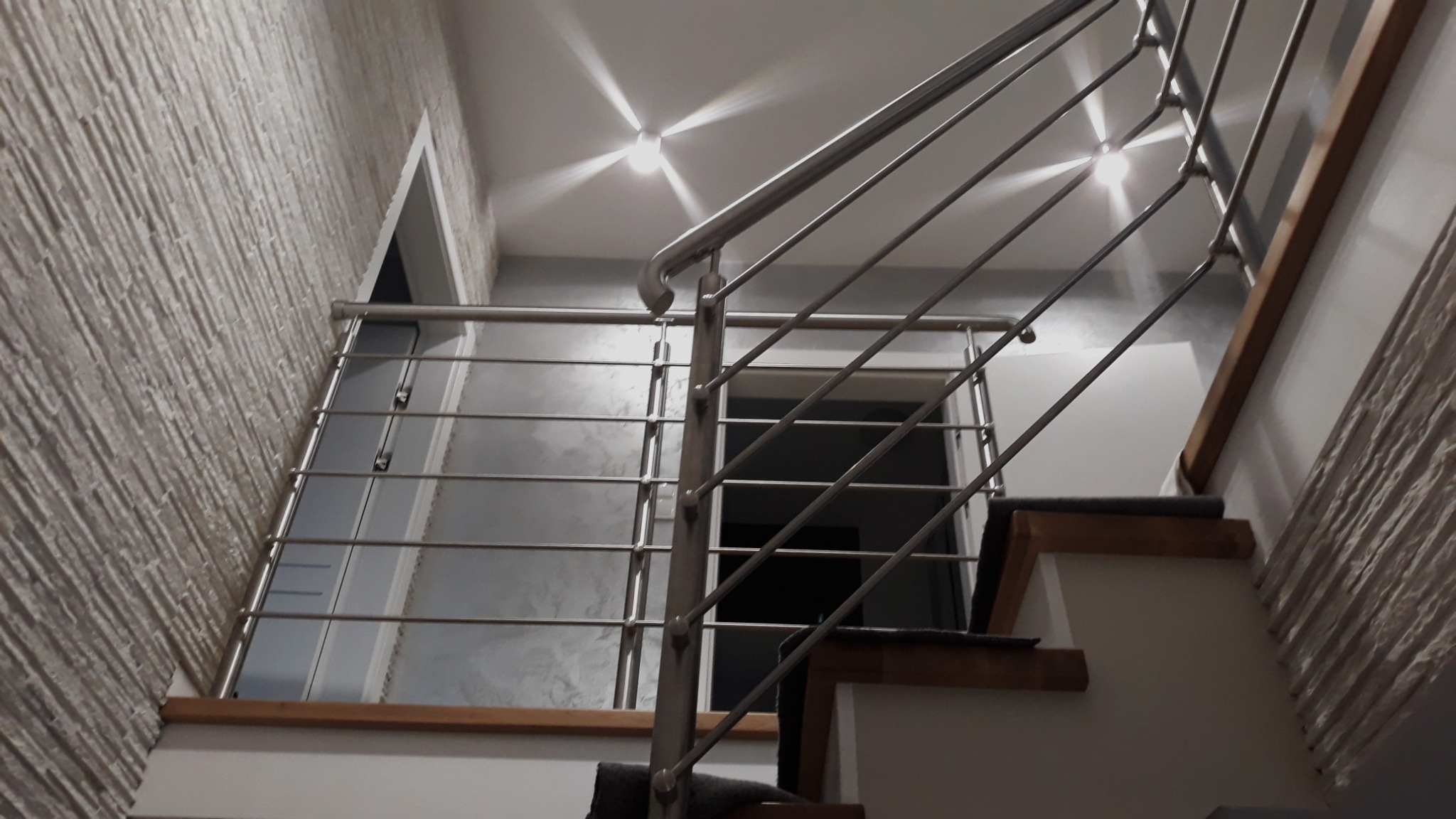 Nowoczesne schody z balustradą ze stali nierdzewnej, widok od dołu, z oświetleniem punktowym na suficie i teksturą ściany.