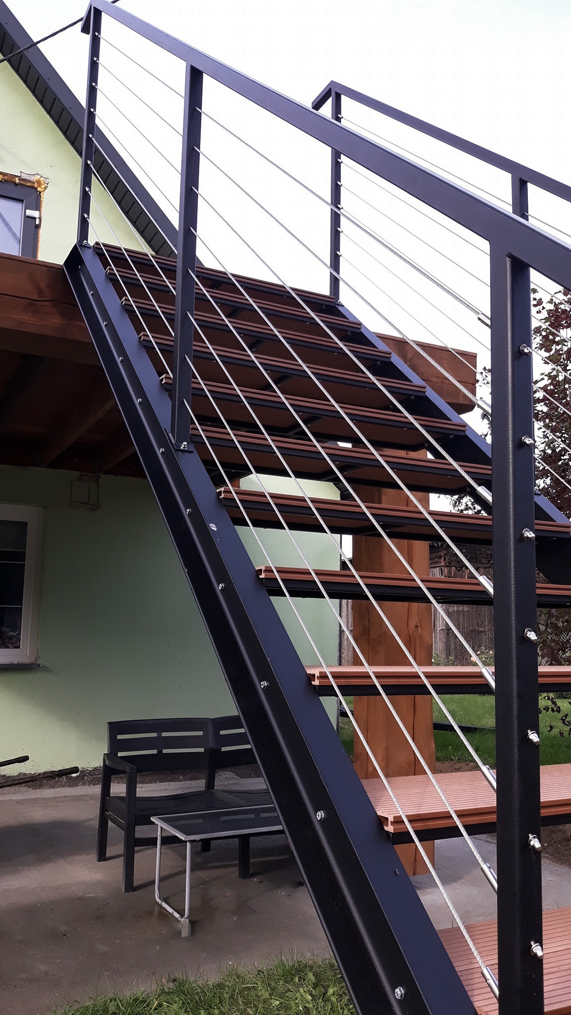 Zewnętrzne schody metalowe z czarną konstrukcją i poziomymi linkami ze stali nierdzewnej, prowadzące na drewniany taras, z widocznym fragmentem elewacji domu w tle.