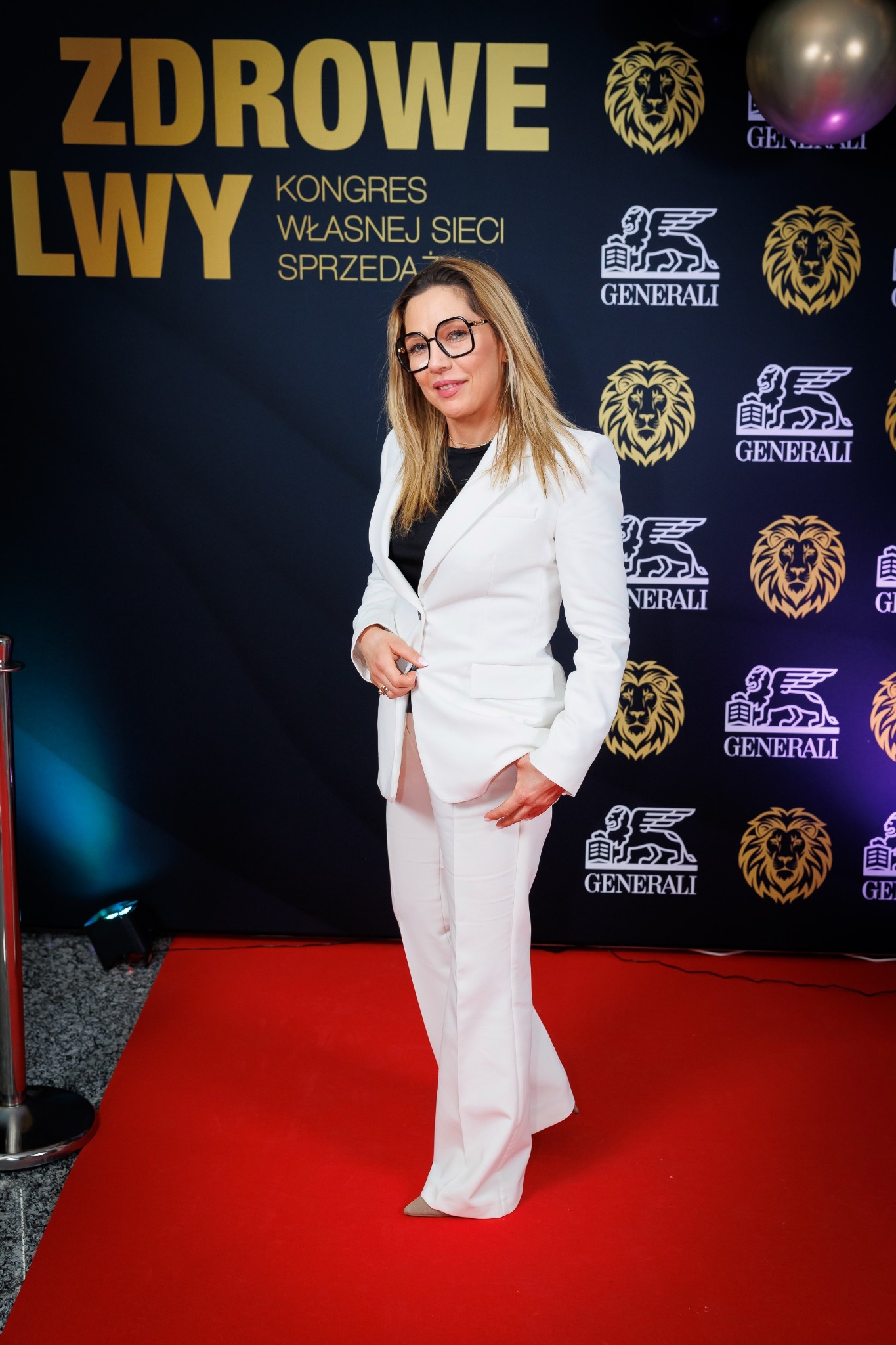 Kobieta w białym garniturze pozuje na czerwonym dywanie przed ścianką z logo firmy ubezpieczeniowej Generali, na tle napisu 'Zdrowe Lwy' i złotego balonu.