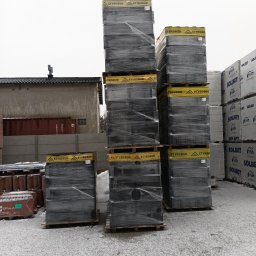 E-IWAN - Stosy palet z zapakowanym styropianem Styrobud i Solbet na placu budowy pokrytym śniegiem, w tle budynek i kontener.