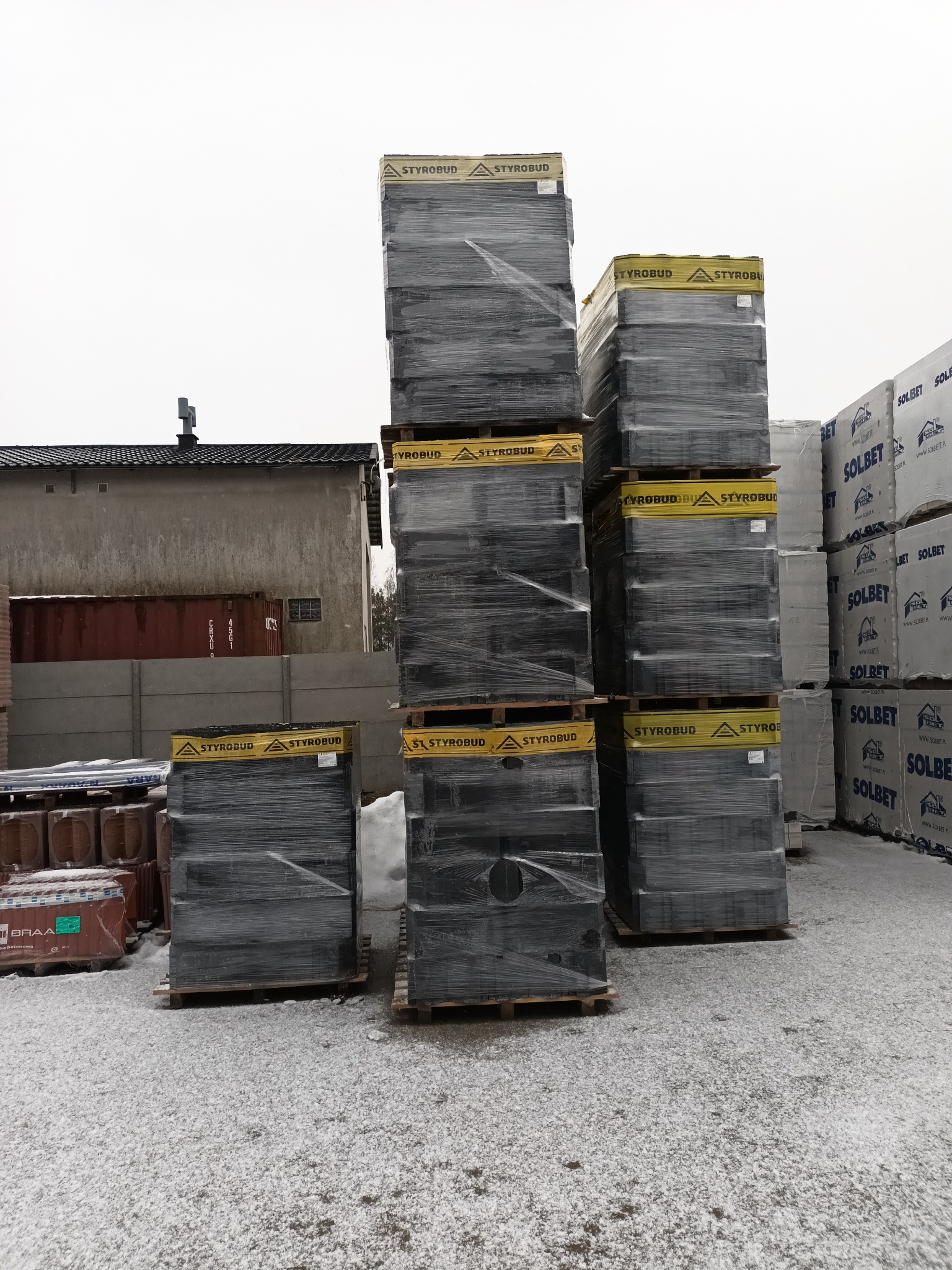 Stosy palet z zapakowanym styropianem Styrobud i Solbet na placu budowy pokrytym śniegiem, w tle budynek i kontener.