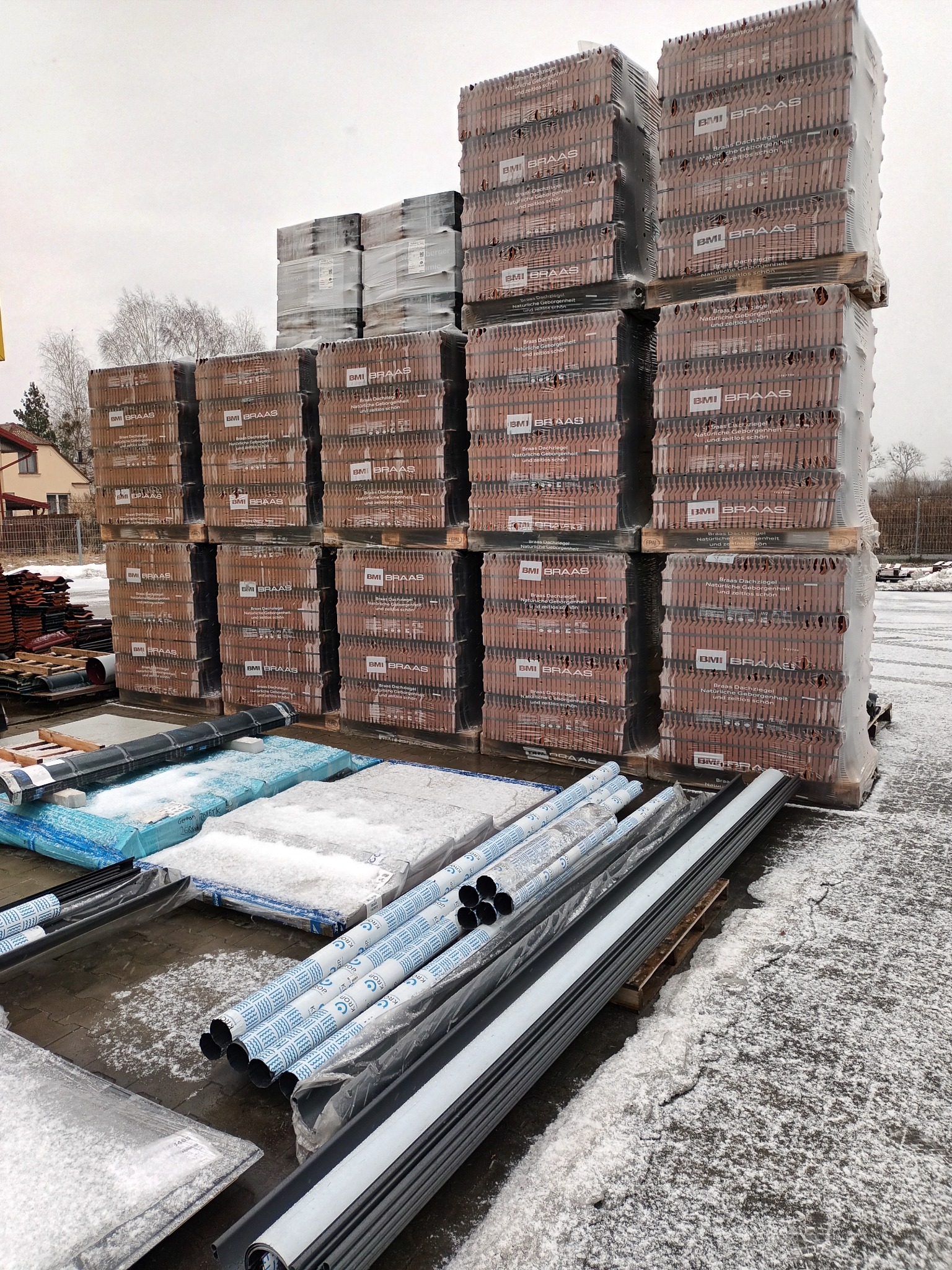 Palety z dachówką ceramiczną marki BMI Braas, folie dachowe i rynny plastikowe na placu budowy w zimowy dzień.