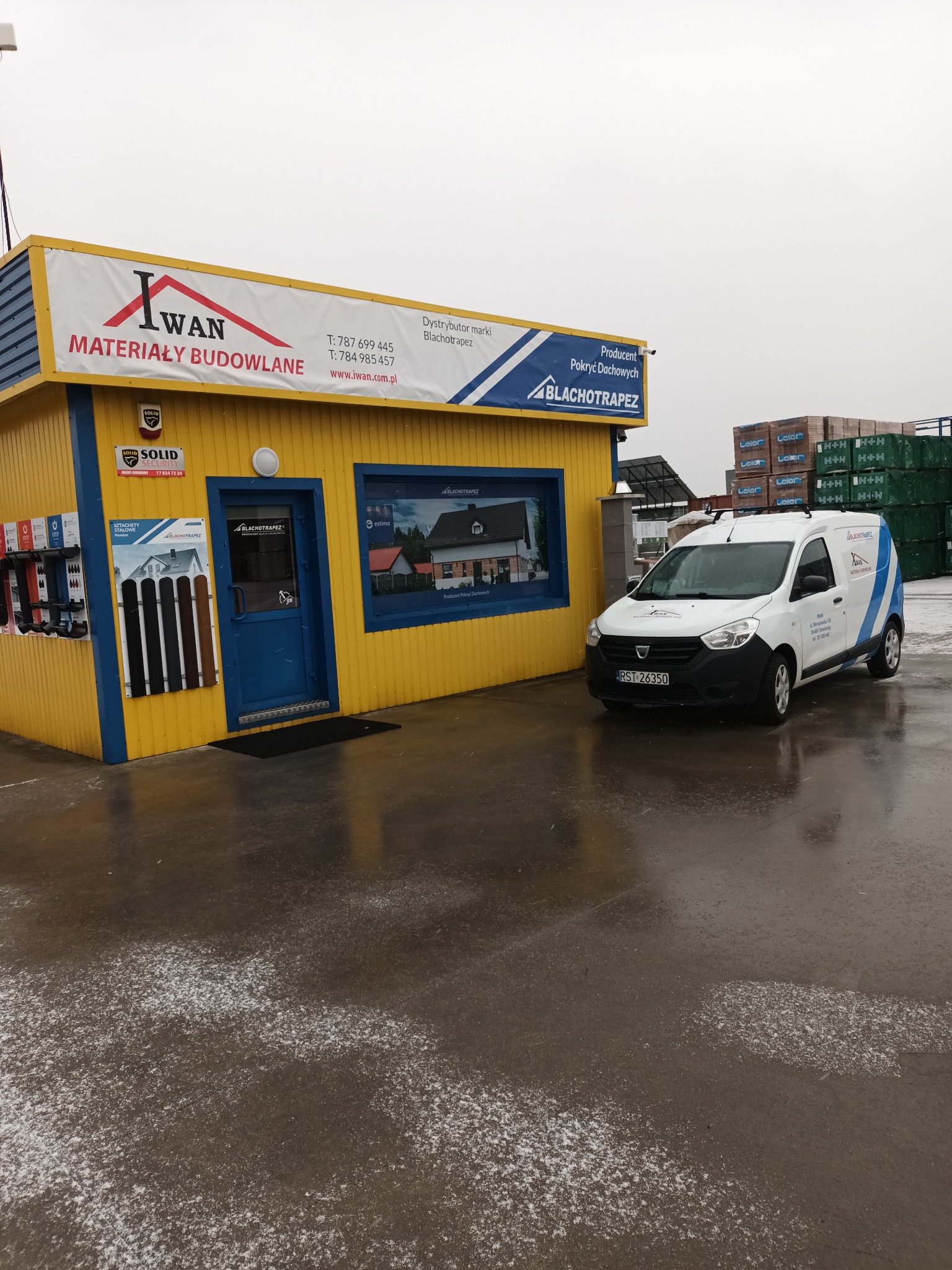 Żółto-niebieski budynek składu budowlanego Iwan z logo w kształcie dachu, obok zaparkowany biały samochód dostawczy z logo Blachotrapez, w tle palety z materiałami budowlanymi, mokra nawierzchnia...