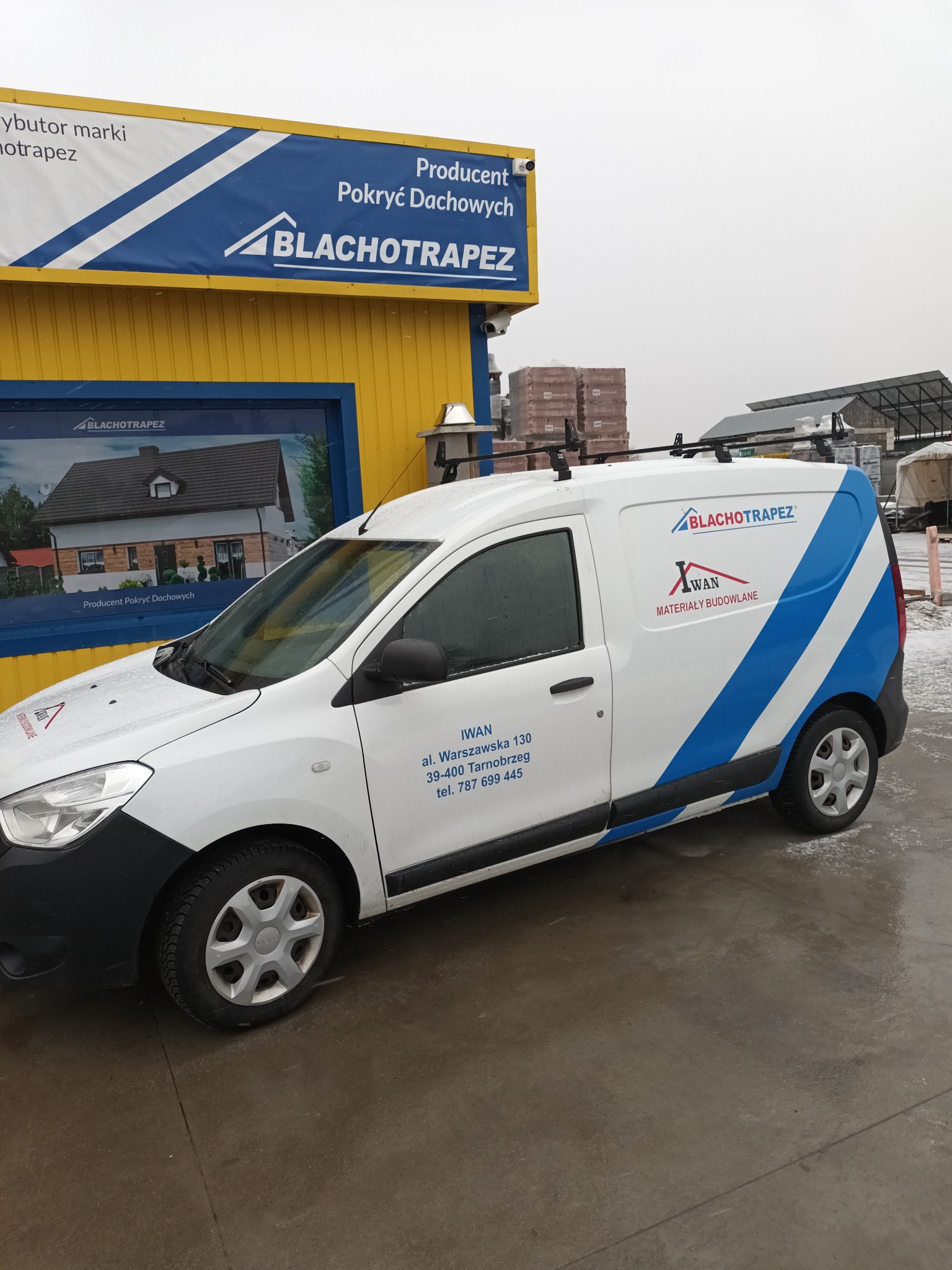 Biała furgonetka z logo Blachotrapez i Iwan Materiały Budowlane zaparkowana przed żółtym budynkiem z ekspozycją dachówek, w tle widoczne składowane materiały budowlane.